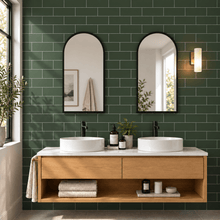 A modern double-sink bathroom vanity with a light wood floating cabinet, white vessel sinks, black faucets, and two arched mirrors set against a dark green subway tile wall.