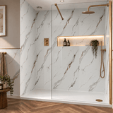 A modern walk-in shower enclosure with a glass door and gold-tone hardware, set in a bathroom with white marble-look walls, a built-in shelf with warm lighting, wood flooring, and small decorative plants.