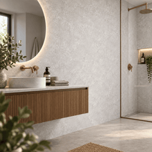 A modern bathroom interior with a floating wooden vanity, round vessel sink, warm brass fixtures, a large oval mirror, and a glass shower enclosure. The wall and floor have a light textured stone or plaster finish, with small plants and bottles placed on the counter.