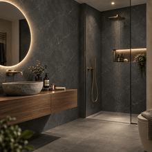 Modern bathroom interior with dark gray stone walls and floor, a floating wood vanity with a stone vessel sink, a round backlit mirror, and a glass-enclosed shower area with gold fixtures.